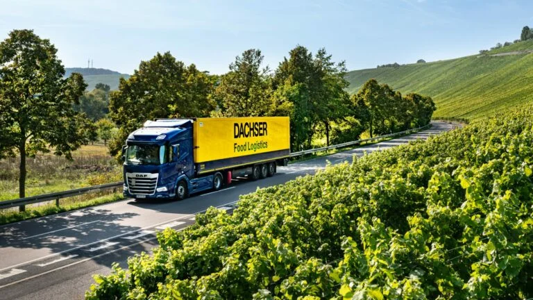 Dachser Food Logistics Lkw fährt durch eine ländliche Landschaft mit Weinbergen am Straßenrand. DACHSER Food Logistics truck drives through a rural landscape with vineyards along the roadside.
