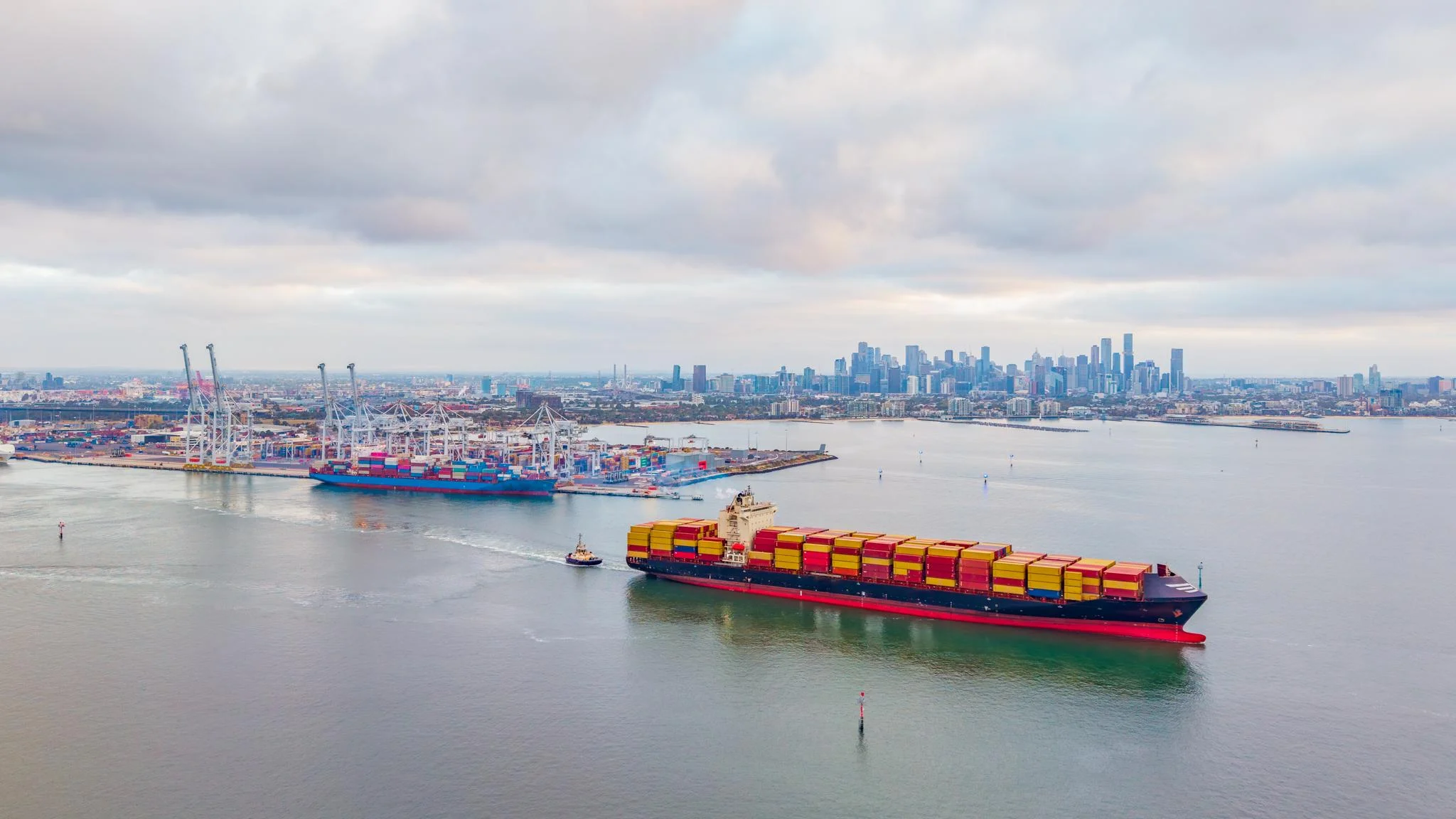 Containerschiff mit Schlepper, das durch Melbournes geschäftigen Hafen fährt, mit Stadtsilhouette, Industriekränen und Frachtterminals unter bewölktem Himmel. – Stockfoto
