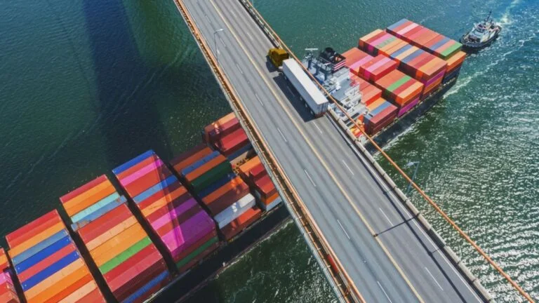 DGI_039601_edit Weiß-gelber Lkw fährt auf einer Brücke, unter der ein Containerschiff fährt